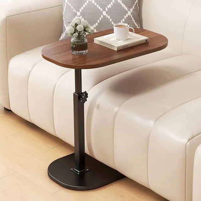 SUSUZI Height Adjustable C-Shaped End Table with a 16"x12" MDF tabletop in a wood finish and a sturdy black metal frame. The table is positioned beside a beige sofa, showcasing its space-saving C-shaped design that slides under furniture. It features an adjustable height mechanism ranging from 23.5" to 31.5" and a lower storage shelf for added functionality. Ideal for use as a sofa side table, laptop stand, or bedside organizer.