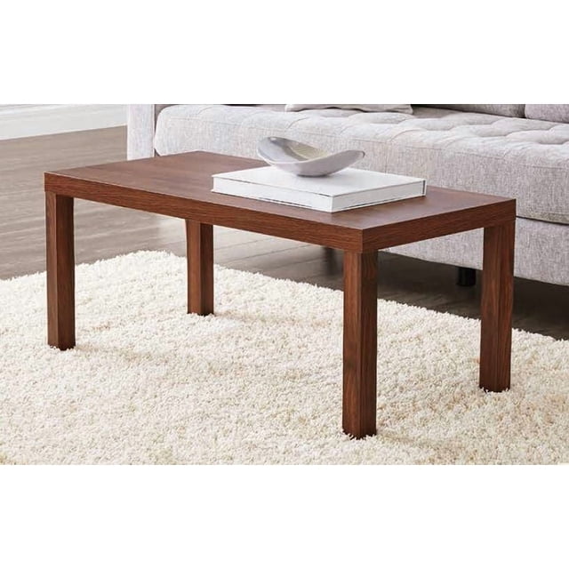 Hometrends Coffee Table in canyon walnut finish with a simple modern rectangular design, four straight legs, and a smooth wood grain surface, placed on a white shag rug in front of a light grey tufted sofa, holding a white book and a decorative bowl, suitable for versatile living room use.