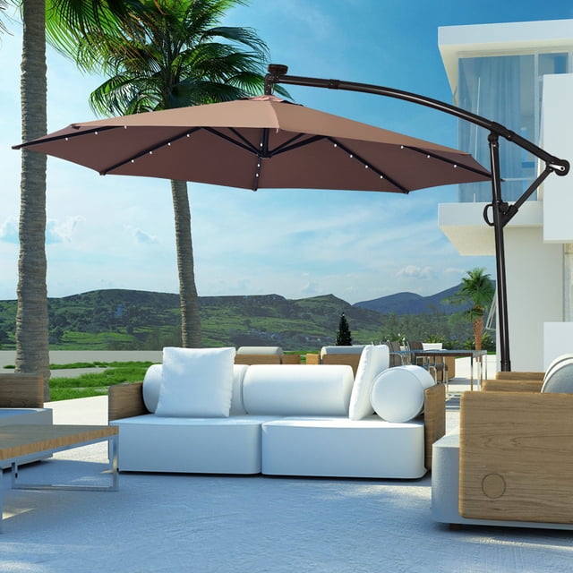 Costway 10FT Patio Offset Umbrella Solar LED 360degrees Rotation Brown displayed in an outdoor setting. The umbrella features a large 10-foot brown polyester canopy supported by a powder-coated aluminum pole with 8 steel ribs. Equipped with 24 solar-powered LED lights along the ribs, it provides illumination at night. The umbrella includes a vented top for airflow and offers 360-degree rotation and tilt adjustment for optimal shade. Positioned near modern patio furniture with a scenic background of palm trees and mountains.