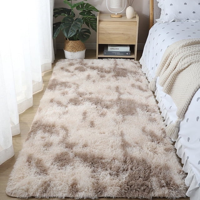 Boovnll Rugs for Bedroom, a super soft and cozy tie-dyed plush throw rug measuring 3.9x5.2 feet. The rug features a beige and brown tie-dye pattern with a thick, artificial fleece surface and high-density sponge interlayer for added comfort. It has a non-slip bottom for stability and is suitable for living rooms, bedrooms, and other home decor settings. Ideal as a runner rug or area rug, it is shown beside a bed in a bright, modern bedroom with wooden furniture and a potted plant.