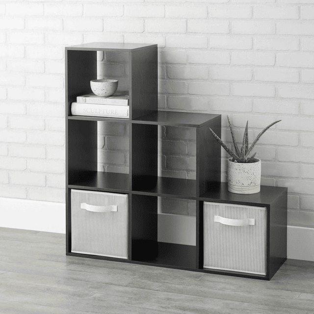 Mainstays 3-2-1 Cube Storage Organizer in espresso finish with six open-back cube shelves arranged in a stair step design, featuring two beige fabric storage bins with white handles in the bottom cubes, a potted aloe plant on the top right shelf, and stacked books with a decorative bowl on the top left shelf, set against a white brick wall and light wood floor. Suitable for organizing books, decor, and small household items.