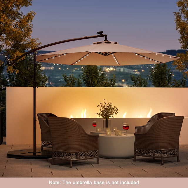 Costway 10 Ft Patio Solar LED Offset Umbrella with 40 lights and crank cross base in tan, ideal for patio shade and outdoor gatherings. The umbrella features a wide 10-foot canopy with solar-powered LED lights along the ribs, providing illumination for nighttime use. Its sturdy steel frame and fade-resistant polyester fabric ensure long-lasting durability, while the easy-tilt adjustment system offers versatile shading options. Displayed in an evening patio setting with wicker chairs and a table, surrounded by ambient lighting. Note: The umbrella base is not included.