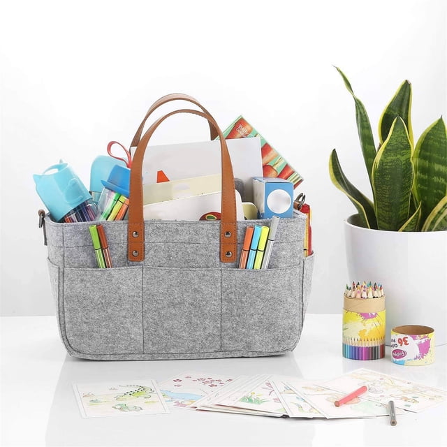 Foldable Diaper Caddy Organizer with Removable Compartments in grey fabric, featuring brown leather handles. The portable baby changing tote is shown filled with various items like markers, books, and baby supplies, demonstrating its multifunctional use. The caddy is placed on a white surface next to a potted plant and art supplies, highlighting its versatility for diapering, travel, or storage needs.