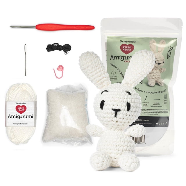 Red Heart™ AMIGURUMI Crochet Collection Kit showing a white crocheted bunny amigurumi with black stitched facial features, accompanied by a red crochet hook, white recycled cotton yarn, polyester stuffing, yarn needle, stitch marker, black thread, and a pattern sheet in resealable packaging, ideal for crafting, gifting, and collectible projects.