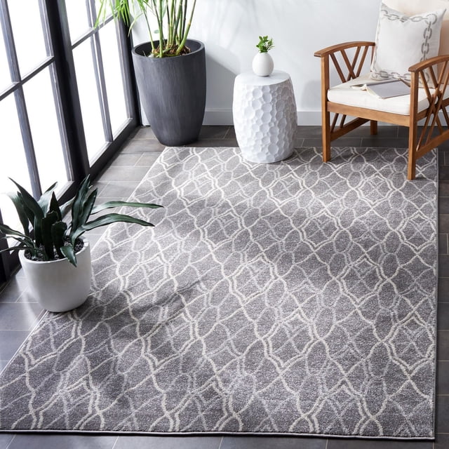 Safavieh Amherst Virginia Geometric Area Rug in grey with white interlocking geometric pattern, power-loomed polypropylene for durability and easy care, placed on a tiled floor near large windows. Styled with a wooden armchair with white cushions, a white textured ceramic side table with a small plant, and two potted plants, ideal for high-traffic indoor or outdoor spaces.
