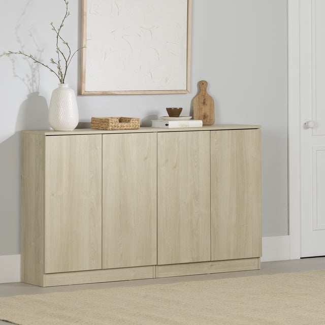 South Shore Arum Set of 2 Sideboards with 2 Doors in bleached oak finish, shown side by side with handleless push-to-open doors and a podium base. The natural wood grain cabinets are topped with a white vase holding branches, a woven basket, a small brown bowl on a book, and a wooden cutting board, set against a light grey wall with framed abstract art.