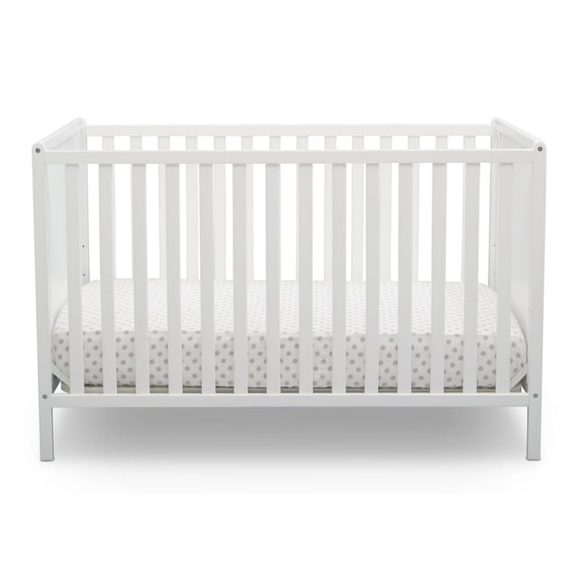 Delta Children Kendrick 4-in-1 Convertible Crib in white with vertical slats and polka dot mattress, featuring adjustable mattress height and sustainable wood, designed as versatile nursery furniture for growing babies.