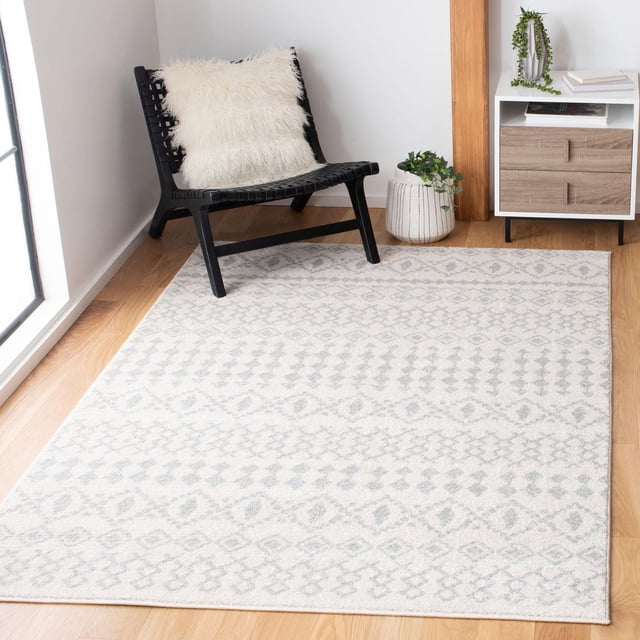 SAFAVIEH Tulum Herbert Distressed Southwestern Area Rug in soft neutral tones with a subtle southwestern geometric pattern, placed on light hardwood flooring in a minimalist room with a black woven lounge chair featuring a white fluffy pillow, a white planter with green foliage, and a white and wood side table. This durable accent rug adds warm, inviting character to living spaces.