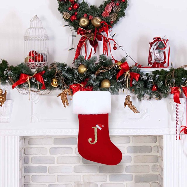 Monogram Christmas Stockings, red velvet stocking with a soft white plush cuff and an embroidered gold letter 'J' adorned with a Santa hat. The stocking is displayed hanging on a white brick fireplace decorated with festive garlands, red ribbons, and gold ornaments, creating a cozy holiday atmosphere.