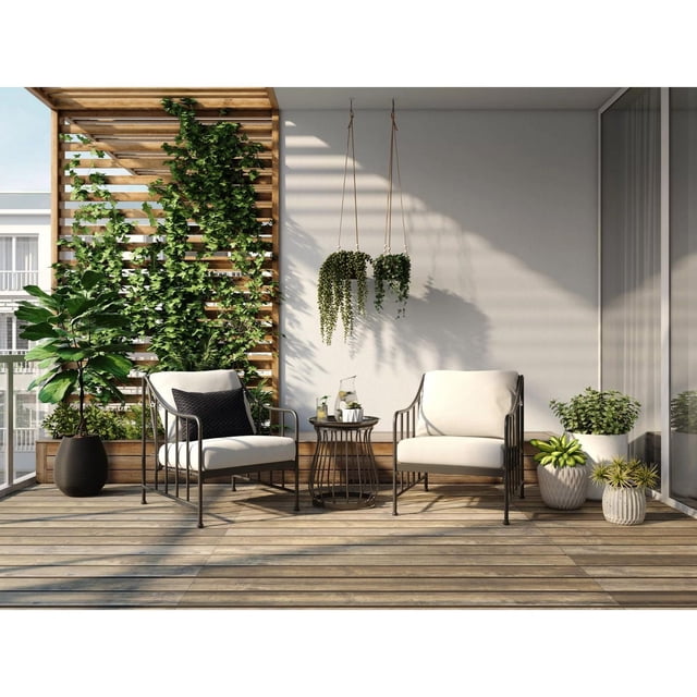 Better Homes & Gardens Aubrey 3-Piece Steel Outdoor Chat Set with Cushions, Cream, featuring two cream cushioned steel frame chairs with black armrests and a small round all-season metal table with a glass pitcher and cups, set on a wooden patio deck with vertical wooden slats supporting climbing plants, two hanging planters, and potted greenery, suitable for outdoor patio conversation and relaxation.