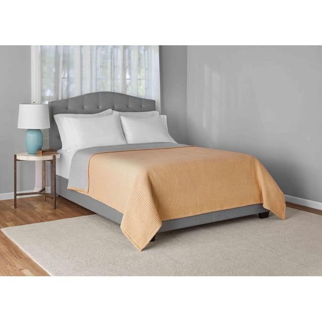 Mainstays Garment Wash Adult Matelasse Quilt in yellow, king size, displayed on a grey upholstered bed with a tufted curved headboard. The quilt features a ribbed cotton texture over white pillows and a grey coverlet in a bedroom with light grey walls, hardwood floors, a beige rug, and a wooden side table with a blue lamp. This machine-washable quilt is ideal for adding warmth and charm to bedrooms.
