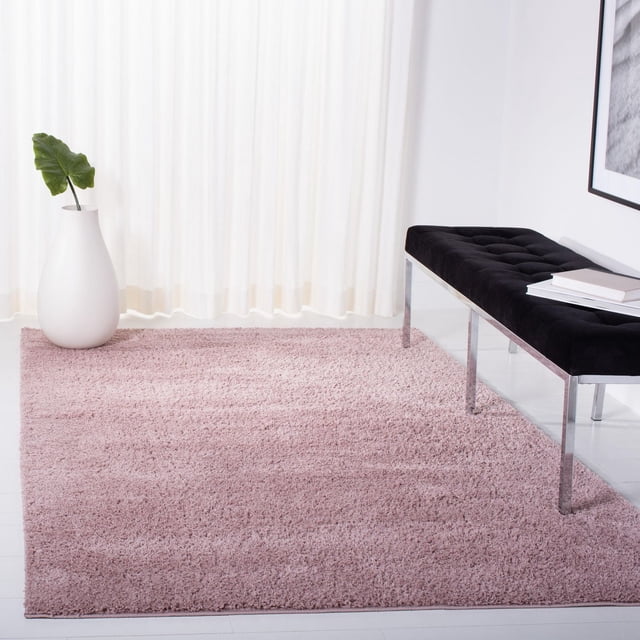 SAFAVIEH August Bronte Solid Plush Shag Area Rug in soft pink, displayed in a bright room with white sheer curtains, a white vase with a green leaf, and a black tufted bench with chrome legs, featuring thick Polypropylène yarn and a Plush texture suitable as a modern accent Rug.