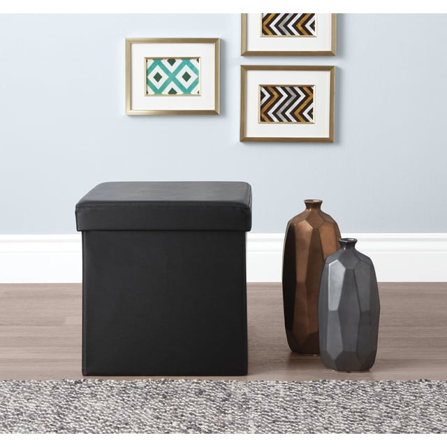 Hometrends 15 inch Storage Cube Black with cushioned lid and foldable design, featuring textured black faux leather exterior, measuring 15 by 15 by 15 inches, placed on wood flooring beside two faceted decorative vases in bronze and gray, beneath three framed geometric artworks on a light gray wall, suitable for storing household items and decluttering spaces.