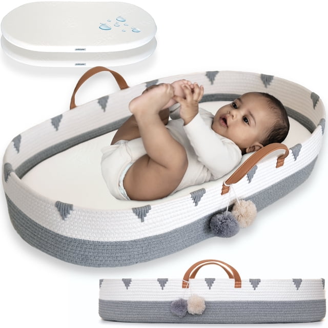 Vesta Baby Cotton Rope Portable Moses Baby Changing Basket Set in gray and white with boho design. The basket features vegan leather handles, pom-pom accents, and a soft foam pad with a waterproof bamboo jacquard cover. A baby is lying comfortably inside the basket, which is suitable for diaper changes or playtime. Includes an extra removable pad cover for convenience.