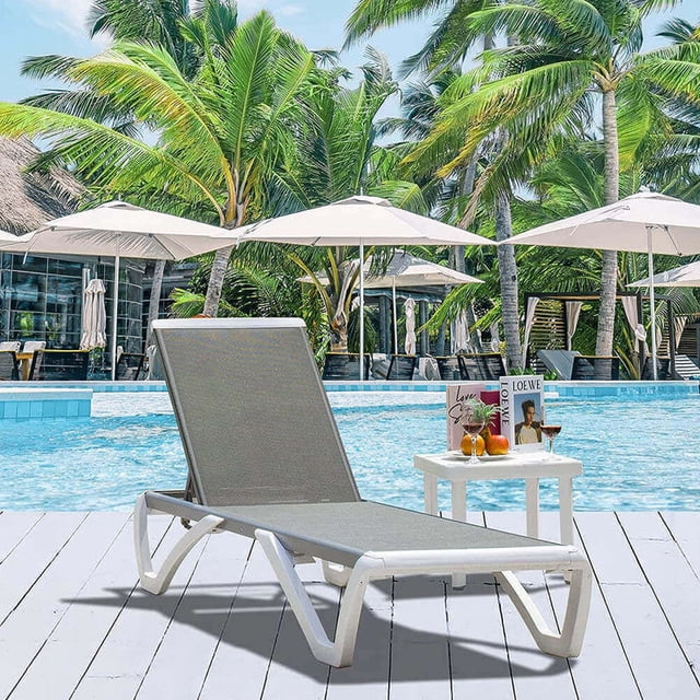 Domi Outdoor Living Aluminum Chaise Lounge Chair with Adjustable Backrest and Side Table, shown in gray, positioned poolside on a white deck. This outdoor lounge chair features a rust-resistant aluminum frame and UV-resistant Textilene fabric for durability and comfort. A small white table accompanies the chair, holding books and drinks, with palm trees and umbrellas in the background, ideal for patio furniture setups.