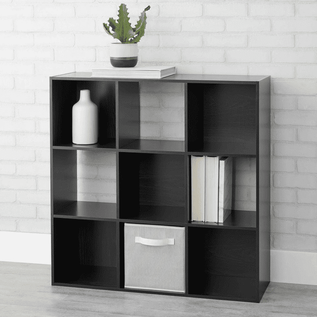 Mainstays 9-Cube Organizer in matte black with nine 11-inch cube shelves, featuring four open-back and five closed-back compartments, holding a white vase, stacked books, and a fabric storage bin, topped with a potted cactus, set against a white brick wall on a light wood floor, suitable for organizing books, decor, and craft supplies.