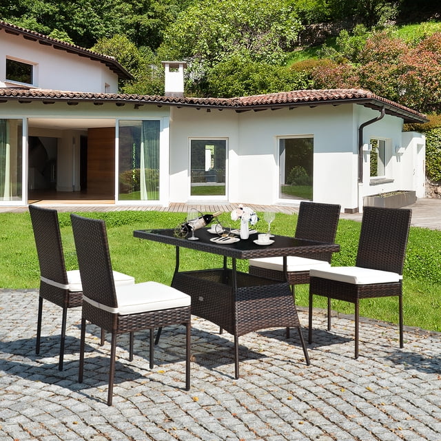 Gymax Ensemble de Salle à Manger de Patio en Rotin 5 Pcs Table et Chaises Ensemble de Mobilier d'Extérieur avec Coussin. l'Ensemble Comprend une Table Rectangulaire en Verre Trempé et Quatre Chaises en Rotin avec Coussins Blancs, Conçu pour l'Usage Extérieur. Fait de Rotin PE durable et d'Acier Enduit de Poudre, le Mobilier Est Résistant aux Intempéries et Robuste. les Chaises Ont une Capacité de Poids de 400 Lb Chacune, et la Table Supporte jusqu'à 180 Lb. les Coussins Sont Amovibles et Ont des Housses en polyester Lavables. l'Ensemble Est Exposé sur un Patio en Pierre dans un Décor de Jardin, avec une Maison Blanche Moderne
