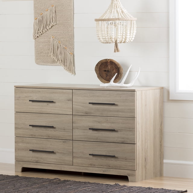 South Shore Primo 6-Drawer Dresser for Adult in rustic oak finish with six large drawers featuring black metal handles and metal slides, designed with contemporary clean lines and a curved kick plate, displayed in a bedroom setting with a woven wall hanging, wooden clock, and white antler decor on top, placed on light hardwood flooring with a dark textured rug nearby.
