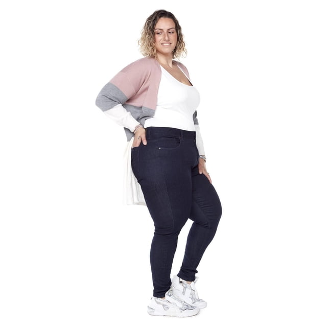 Frankie & Stella ™ Classic Skinny PLUS - Dark Blue jeans worn in a 3/4 profile by a woman paired with a white tank top and a long cardigan featuring pink, gray, and white color blocks. The slim leg jeans are crafted from dark blue super soft flex denim with a high-rise waistband and front zipper, designed for plus sizes with a shape-lift fit. She wears white sneakers with gray accents. These jeans offer flexible movement and are ideal for casual everyday wear.