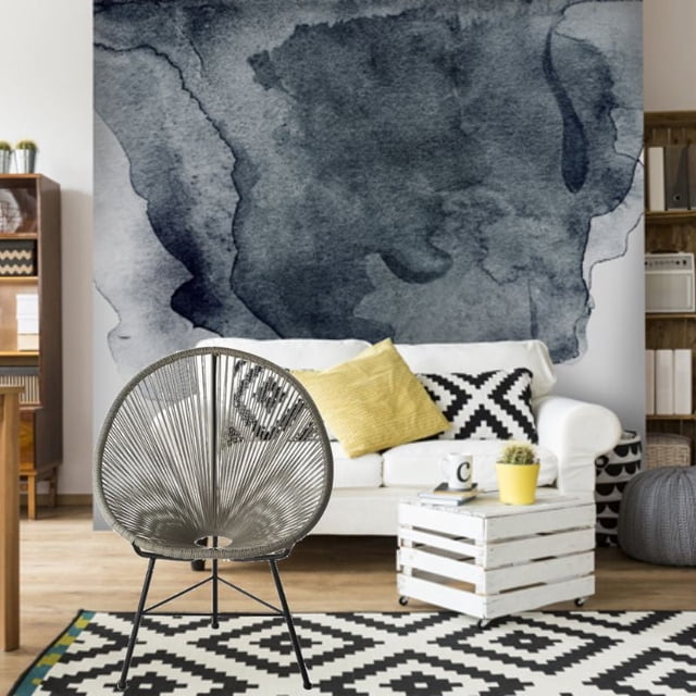 Costa Lounge Chair in grey synthetic rattan with ergonomic oval wire design and high back, positioned indoors with a white sofa featuring yellow and black geometric cushions, a white wooden crate coffee table with a yellow planter, and a large dark blue abstract watercolor wall mural.