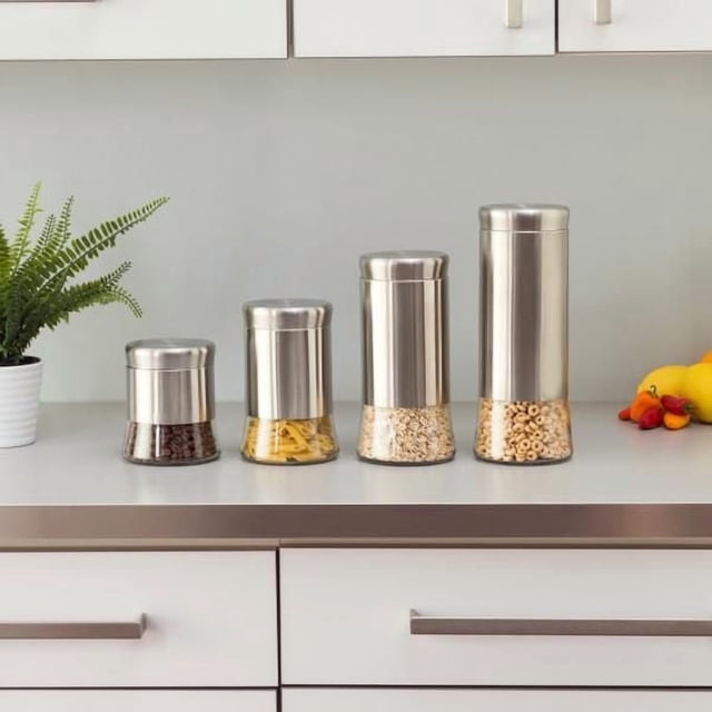 Essence Collection Ensemble de 4 Contenants en Acier Inoxydable avec bases en Verre Transparent Mettant en Valeur les Grains de Café, les Pâtes Sèches, l'Avoine et les Céréales, avec Couvercles Torsadés Hermétiques en Argent Brossé, Disposés sur un Comptoir de Cuisine Blanc avec une Fougère en Pot et un fruit, Idéal pour Organiser des Aliments Secs Ou de Petits Articles Ménagers.