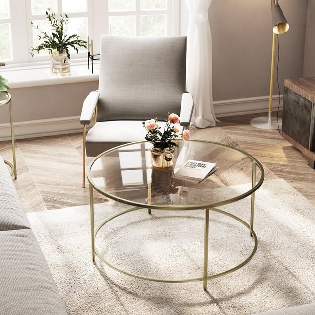 ViscoLogic Regal Mid-Century Round Clear Glass Coffee Table with Metal Frame, Golden. This coffee table features a tempered clear glass tabletop and a gold-coated metal frame, offering a sleek and modern design. The round table is styled in a cozy living room setting with a grey armchair, a white rug, and decorative accents like a potted plant and a book. Ideal for use as a center table or accent piece in modern home décor.
