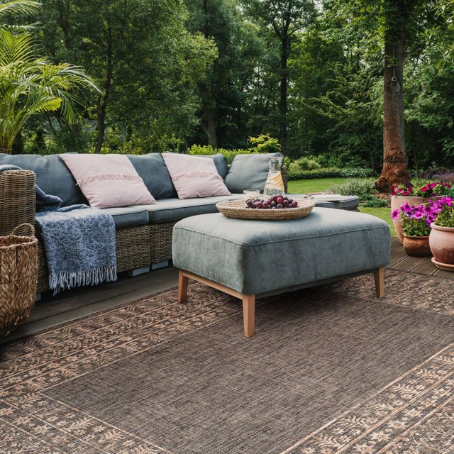 SAFAVIEH Courtyard Jolyon Bordered Indoor/Outdoor Area Rug displayed on a wooden deck patio featuring a brown woven border and intricate pattern. The rug lies under a grey upholstered ottoman with wooden legs, beside a wicker sectional sofa with blue cushions and pink pillows. Surrounded by green trees and potted purple flowers, this durable polypropylene rug suits both indoor and outdoor use and is easy to clean with a garden hose.