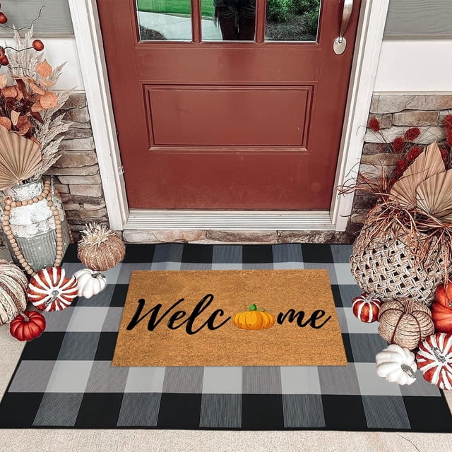 Cotton Plaid Rug in an orange buffalo plaid design, styled as a fall outdoor front door decorative mat. The hand-woven rug is layered under a 'Welcome' doormat with a pumpkin graphic, placed on a porch surrounded by autumn-themed decor, including pumpkins and dried plants. The rug measures 35.4 x 23.6 inches and is made of cotton with a low pile and rubber backing, suitable for porches, entryways, and farmhouse settings.