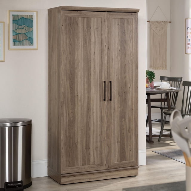 Sauder® HomePlus Armoire de Rangement de la Collection en Fini Bois Brun Pâle avec Deux Portes à Panneaux Encadrées et Poignées Verticales Noires, Posée sur un Plancher de Bois Clair à Côté d'Une Poubelle en Acier Inoxydable. Cette Armoire de Plancher Offre un Rangement Polyvalent Idéal pour le Mobilier de Salle de Bain, le Garde-Manger Ou l'Organisation de la Salle Utilitaire.
