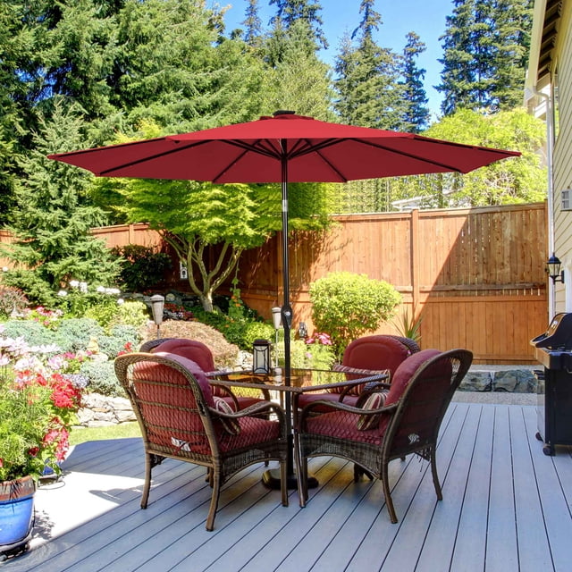 Costway Parasol de Marché pour Patio de 10 Pieds à DEL Solaires en Bourgogne avec Réglage d'Inclinaison et Manivelle, Installé sur un Patio en Bois Entouré de Verdure Luxuriante et d'Une Clôture en Bois. le Parasol Présente une Auvent en polyester de 10 Pieds Soutenu par un Poteau en Fer de 1,5 Po et 8 Nervures Robustes. Il Comprend 24 Lumières DEL à Énergie Solaire pour l'Éclairage Nocturne et un Auvent Ventilé pour une Meilleure circulation de l'Air. l'Ensemble Comprend une table Ronde avec Quatre Chaises en Osier sous le Parasol