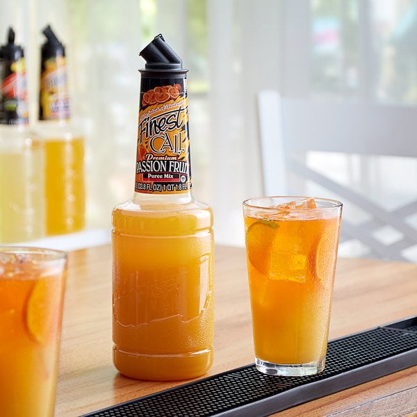 Finest Call Bouteille de Mélange de Purée de Fruit de la Passion Premium de 1 Litre avec Bouchon Verseur Breveté Exposée sur un Comptoir de bar en Bois à Côté d'Un Verre de cocktail de Fruit de la Passion de Couleur Orange Garni de Glaçons et de Tranches de Fruits. Connu pour Sa texture Lisse, Ce Mélange de Purée Est Idéal pour les cocktails, desserts et smoothies. l'Arrière-plan Comprend des Bouteilles Supplémentaires et une Mise en Scène Vive et Aérée.