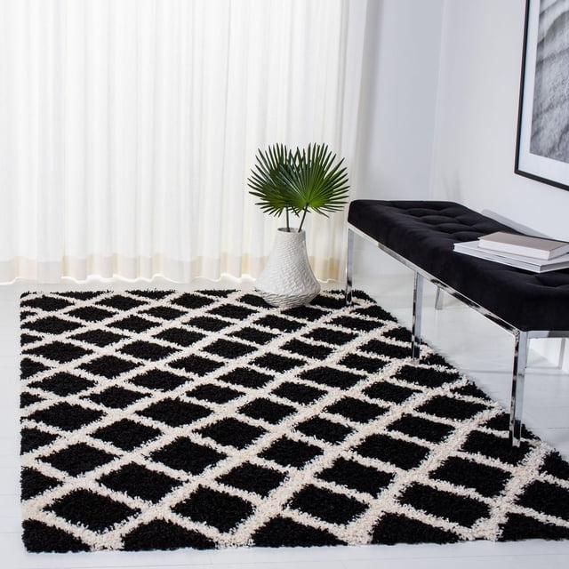 Safavieh Dallas Jerrie Geometric Shag Area Rug with black and white diamond pattern and plush shag texture, placed on white flooring beside a black tufted bench with chrome legs and a white vase holding green palm leaves, in a bright room with sheer white curtains. This durable accent rug is ideal for high-traffic areas and adds texture to contemporary spaces.