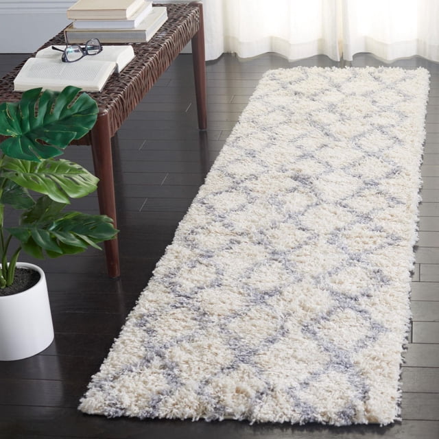 Safavieh Venus Nanda Geometric Shag Area Rug with a 1.77-inch medium pile in cream and gray geometric diamond pattern on dark hardwood floor, styled beside a woven bench with books and a potted green plant, suitable for adding plush texture and warmth to modern or boho interiors.