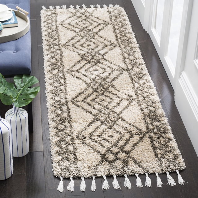 Safavieh Moroccan Fringe Erksine Geometric Shag Area Rug runner featuring cream and dark brown tribal geometric patterns with plush synthetic yarn texture and white fringe tassels on both ends, placed on dark hardwood floor near a blue bench and decorative vases, ideal for adding comfort and style to narrow hallways or entryways.