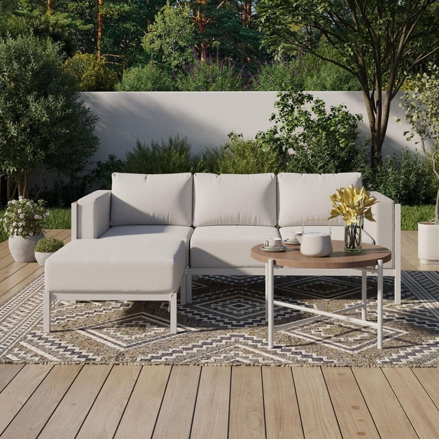 HOMETRENDS Westmont 4-Piece Steel Outdoor Sectional Set with Cushions & Covers, White, featuring a white powder-coated steel frame sectional with three cushioned seats and chaise lounge, paired with a round steel tabletop coffee table with woodgrain finish on a geometric patterned outdoor rug, set on a wooden patio deck surrounded by green shrubs and trees, suitable for enhancing outdoor living and patio furniture spaces.