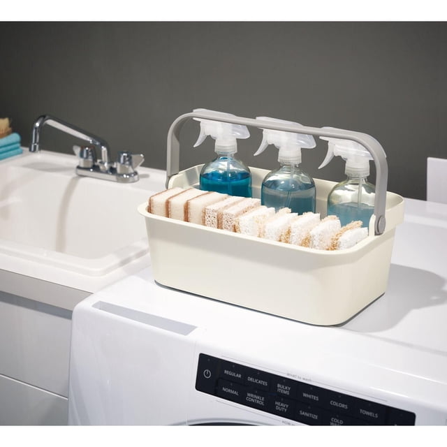 iDesign Coconut & Gray Eco BPA-Free Recycled Plastic Stackable Open-Front Large Basket with Handles measuring 12.9" x 8.7" x 5.1", holding three spray bottles with blue liquid and multiple cleaning sponges, placed on a white washing machine next to a sink with chrome faucet, suitable for organizing cleaning supplies in laundry or bathroom areas