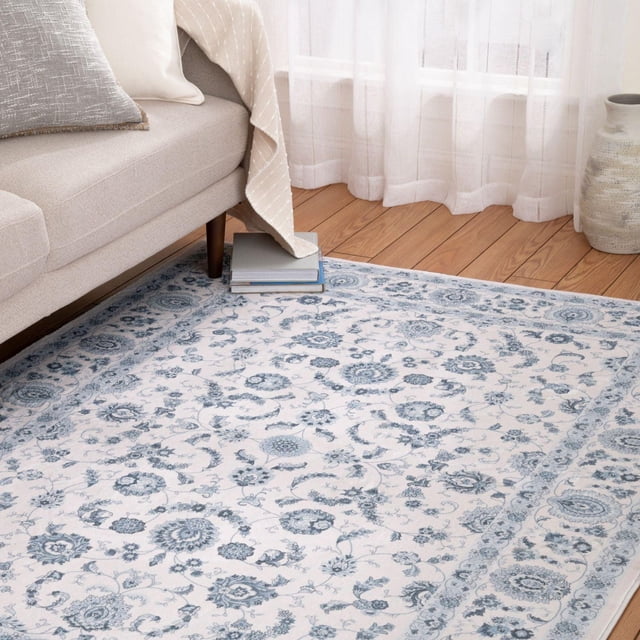 Banburry Ivory and Grey Woven Area Rug with intricate blue and grey floral patterns on an ivory background, placed on hardwood flooring beside a beige sofa with a grey pillow and beige throw, near sheer white curtains and a decorative ceramic vase, suitable for enhancing living room or home office decor.