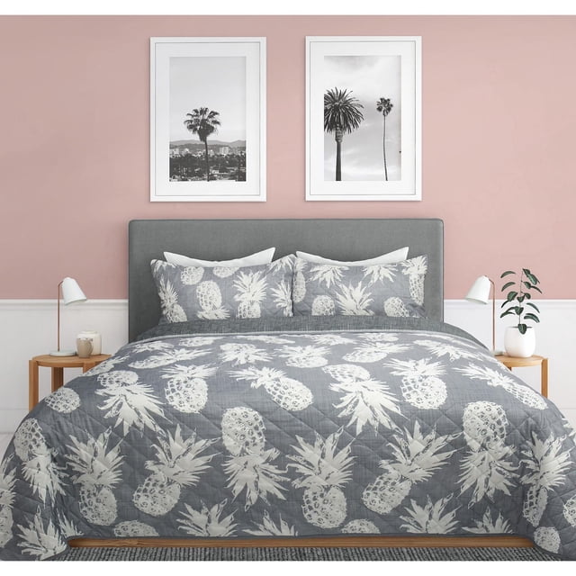 Safdie & Co. Quilt Set 3PC K Pineapples with a grey microfiber quilt and two matching shams featuring white pineapple prints, displayed on a bed with a grey upholstered headboard against a pink wall with two black-and-white palm tree photos, flanked by wooden nightstands with white lamps and a small plant, ideal for adding comfort and style to any bedroom.