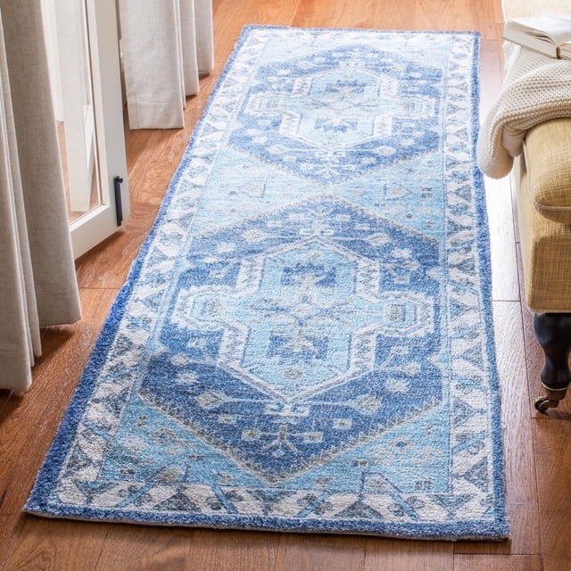 Safavieh Saffron Magdalena Bordered Oriental Area Rug in blue and white geometric medallion pattern, handmade from soft cotton, placed on a hardwood floor beside beige curtains and a tan chair with a knitted throw and open book, ideal as an accent rug for home decor.