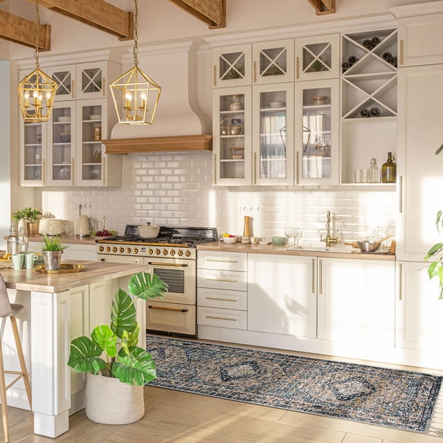 ECARPET Tapis de Style Vintage Traditionnel pour Couloirs, Chambre et Salon Collection Bolivia Bijar avec un Motif Vintage Bleu et beige Atténué, Placé sur un Plancher de Bois Clair dans une Cuisine avec des Armoires Blanches avec des Façades en Verre, des Comptoirs en Bois et un Poêle beige avec des Boutons Dorés. le Tapis Se Situe sous un Îlot de Cuisine avec une Plante Verte en Pot, Illuminé par Deux Suspensions Géométriques Dorées Suspendues au Plafond avec des Poutres en Bois Exposées.