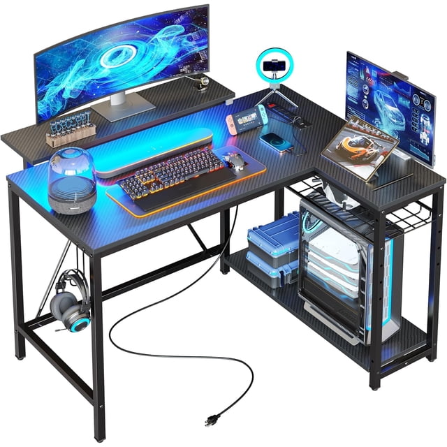 Bestier 42'' Small L Shaped Gaming Desk with Power Outlets in carbon fiber black. This desk features a durable carbon fiber finish, adjustable RGB LED strip lights, and a removable monitor stand that can be installed on the front or side. It includes a built-in charging station with 3 AC outlets and 2 USB ports for powering laptops, gaming gear, and other devices. The desk has reversible 3-tier storage shelves for books, files, or gaming accessories, and adjustable feet for stability. Made with P2 particle board and a metal frame, it is scratch-resistant and easy to clean. Displayed in a gaming setup with monitors, a keyboard, and other accessories.
