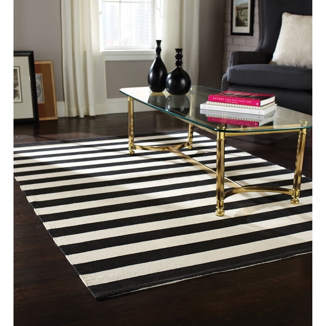 Home Trends Area Rug 4 Ft. 11 In. X 6 Ft. 9 In. Black/White Stripe made of natural fibers with a flat weave texture, stain-resistant and easy to clean. The rug lies on dark hardwood flooring under a glass coffee table with gold metal legs, decorated with black vases and a stack of books. Suitable for adding a modern accent to living rooms.