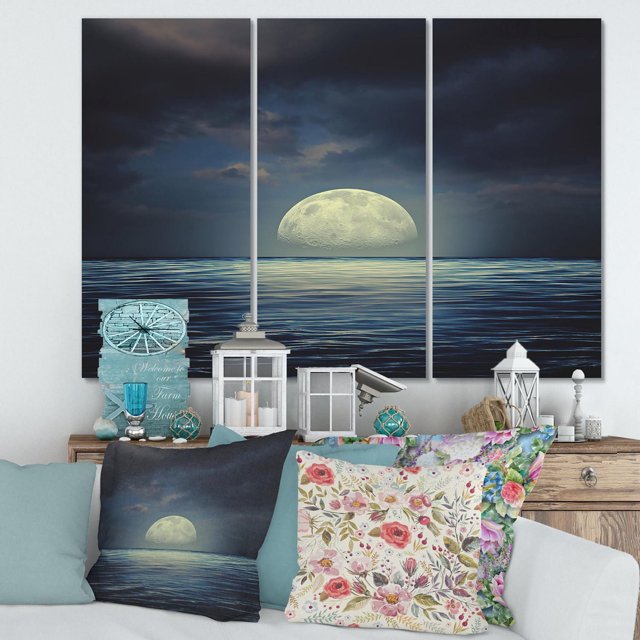 Designart Super Moon Over The Sea II Canvas Wall Art is a three-panel gallery-wrapped cotton canvas print depicting a large moon rising over a calm blue sea under dark clouds, displayed above a wooden console with nautical decor and floral cushions on a white sofa, arriving ready to hang.