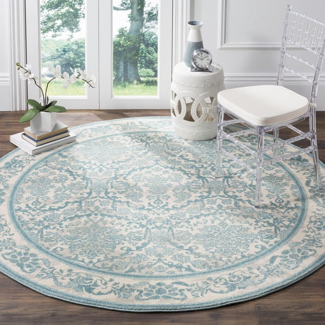 Safavieh Evoke Lorna Traditional Area Rug, round with a soft blue and cream floral pattern in power-loomed polypropylene frieze, placed on hardwood flooring near glass doors. The scene includes a clear acrylic chair with white cushion, a white ceramic garden stool with decorative cutouts, a blue and white vase, a silver clock, and a stack of books topped with a white orchid plant. This durable accent rug is often used to enhance comfort and style in living rooms or reading nooks.