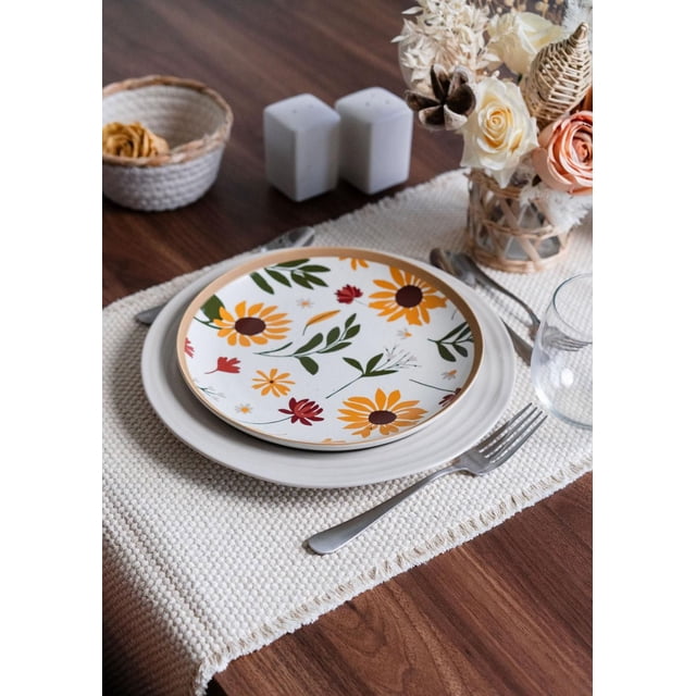 A close-up of a dining table setting on a dark wood table with a white woven placemat. It includes two stacked ceramic plates: a plain white base plate and a smaller top plate featuring yellow and red floral patterns with green leaves. Stainless steel fork and knife are placed beside the plates, along with a clear drinking glass. In the background, a woven basket with dried flowers, white salt and pepper shakers, and a glass vase wrapped in wicker holding dried cream and peach flowers complete the arrangement.
