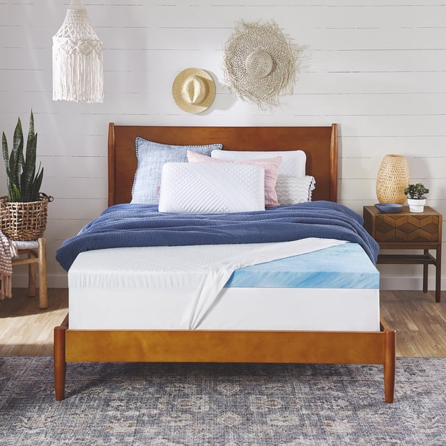 nue by Novaform Gel Memory Foam Mattress Topper, Full, 3-inch, Dual Cooling, displayed on a wooden bed frame in a cozy bedroom setting. The topper features a cool blue gel memory foam layer and a fitted, multi-patterned white cover that stretches to fit mattresses up to 17 inches deep. The bed is styled with blue and white bedding, decorative pillows, and a woven rug, with natural accents like a wicker basket and wall-mounted hats in the background.