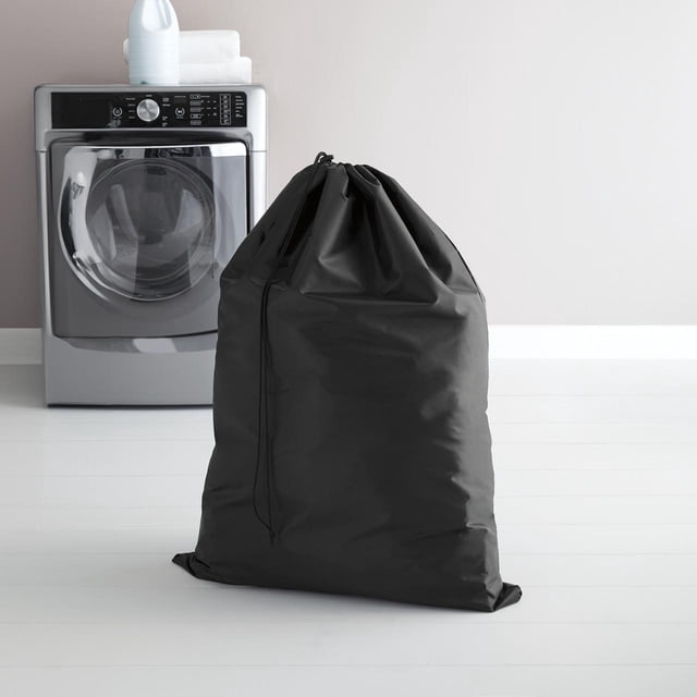 Mainstays Laundry Bag in black with large capacity, durable breathable fabric, and secure drawstring toggle closure, standing on a white floor in front of a silver front-load washing machine with detergent and folded towels on top, suitable for laundry room storage and organization.