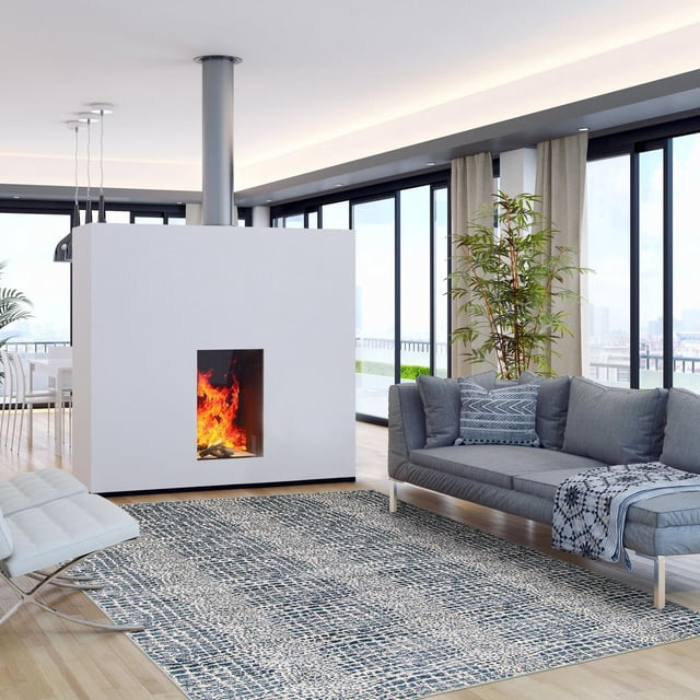 ECARPET Talia Tapis Bleu Marine avec Motif Abstrait Texturé Bleu Marine et beige Placé sur un Plancher de Bois Franc Clair dans un Salon Moderne avec des Fenêtres du Sol au Plafond. Positionné devant un sofa Gris avec des Coussins à Motifs et un Banc Blanc, Près d'Un Foyer Minimaliste Blanc avec des Flammes Visibles et une Grande Plante Verte. le Tapis Est Fabriqué en Fil durable, Idéal pour Rehausser la Déco Intérieure.