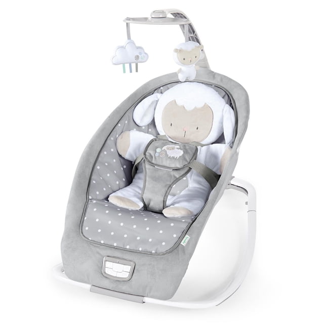 Ingenuity Rocking Seat - Cuddle Lamb in grey, a baby rocker with a soft padded seat featuring white polka dots, a secure harness with a lamb applique, and an overhead mobile with plush lamb and cloud toys. It offers three recline positions and a lightweight frame for easy room-to-room use, suitable from newborn to toddler.