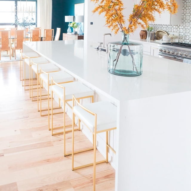 Heavenly Collection Stool with Gold stainless steel frame set of 6, featuring white cushioned seats and slim gold metal legs with 4.25-inch backrests, arranged under a white kitchen island countertop in a bright kitchen with light hardwood flooring and a glass vase with orange flowers on the counter, ideal for kitchen or bar seating.
